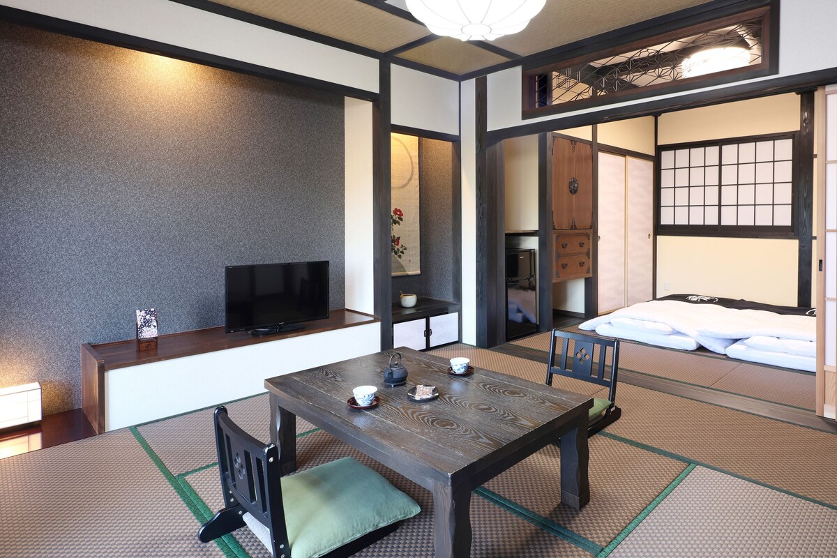 A traditional Japanese-style room with tatami flooring, a low table with cushions, a television, and a sliding door leading to a bedroom.
