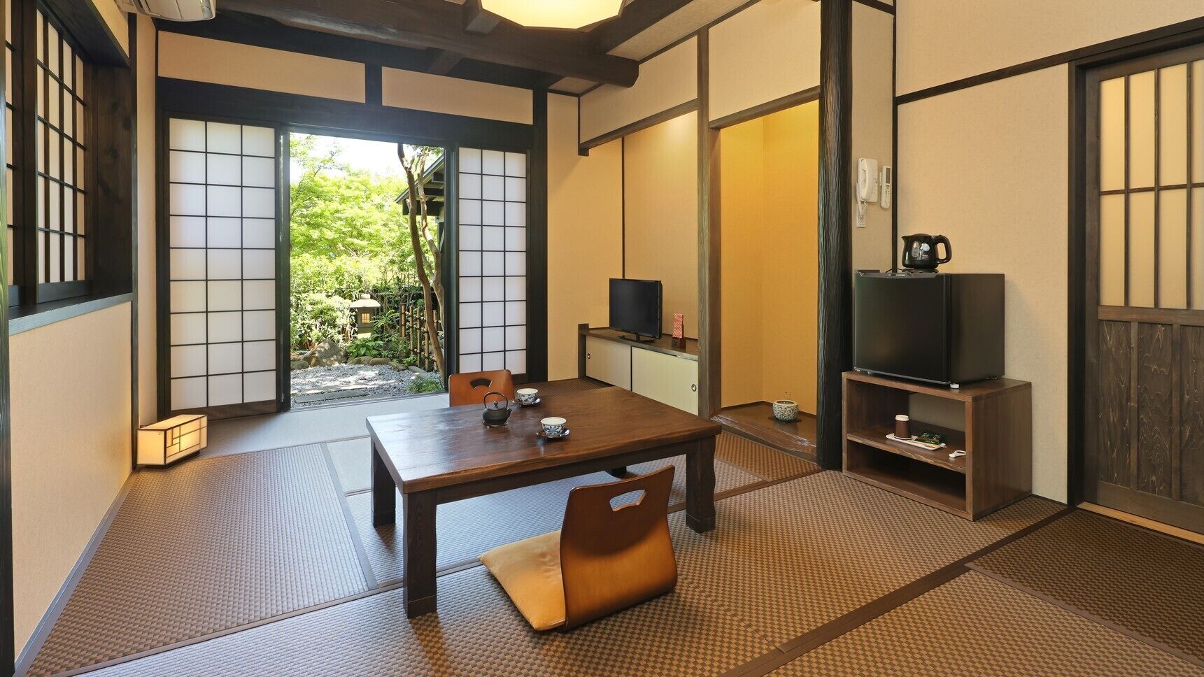 A traditional Japanese room with tatami flooring, a low table, chairs, a television, and a sliding door leading to a garden.