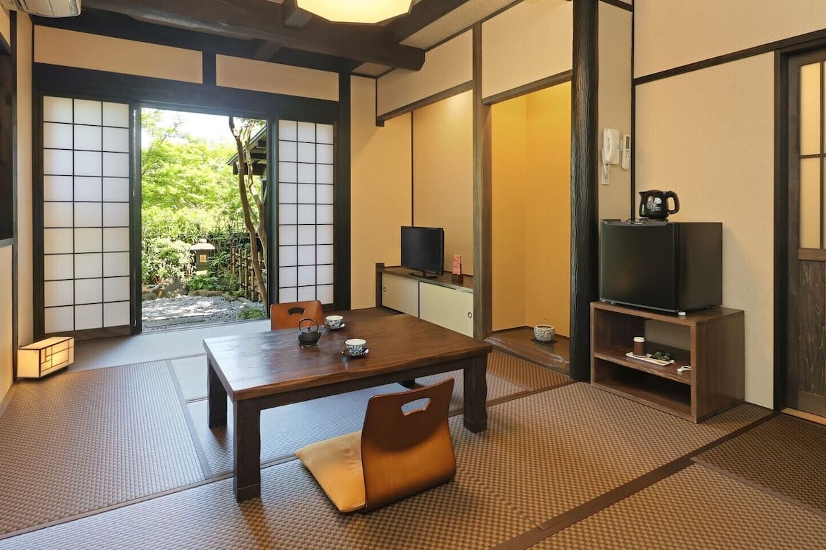 Ein traditioneller japanischer Raum mit Tatami-Bodenbelag, einem niedrigen Tisch, Stühlen, einem Fernseher und einer Schiebetür zu einem Garten.