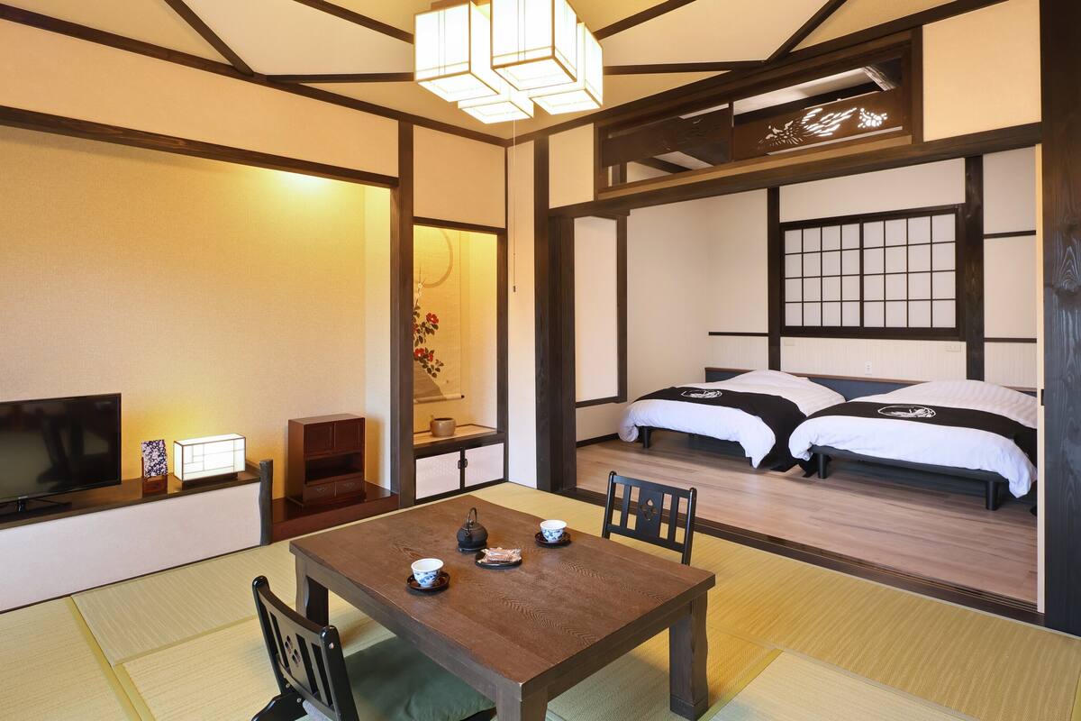 Ein traditioneller japanischer Raum mit Tatami-Boden, einem niedrigen Tisch mit Teeservice, einem Fernseher und einem Bett mit Futonen.