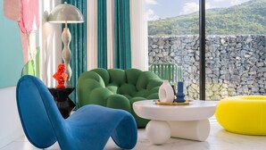 A modern living room with a blue chair, green bean bag, and a yellow bean bag. There is a white table with decorative items and a lamp. The room has a view of a stone wall and greenery outside.