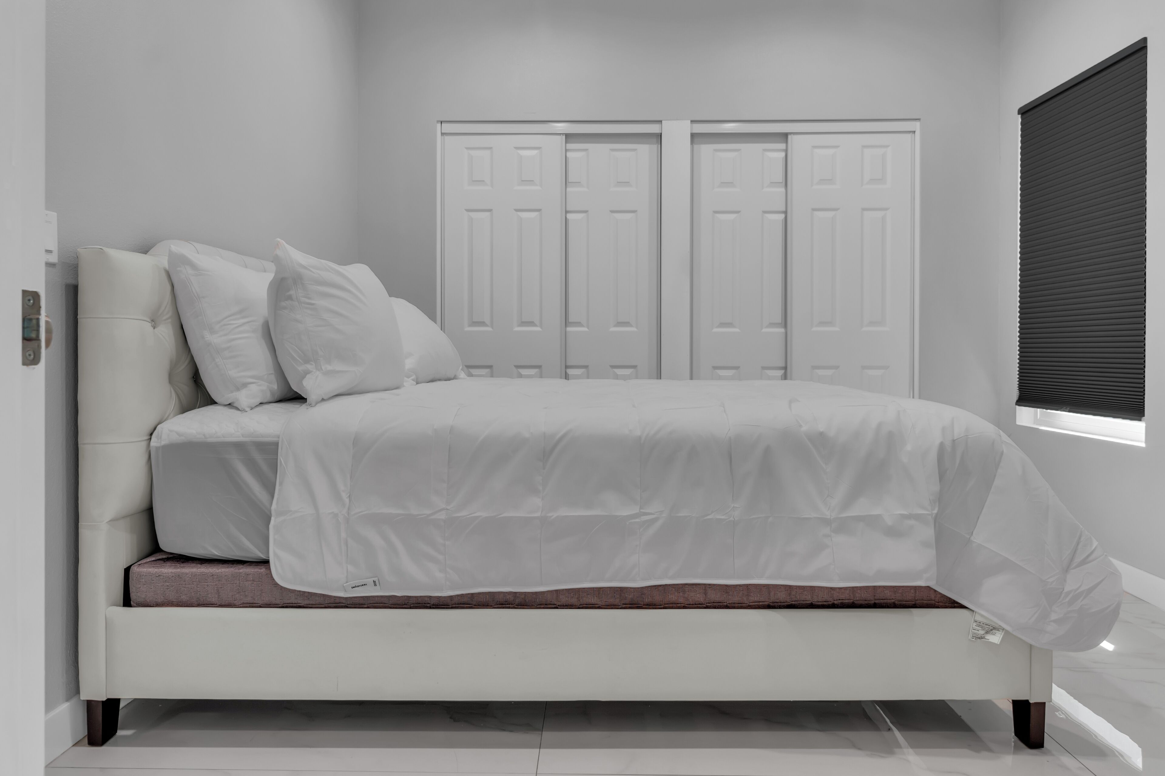 A modern bedroom with a white bed, two doors, and a window with blinds.