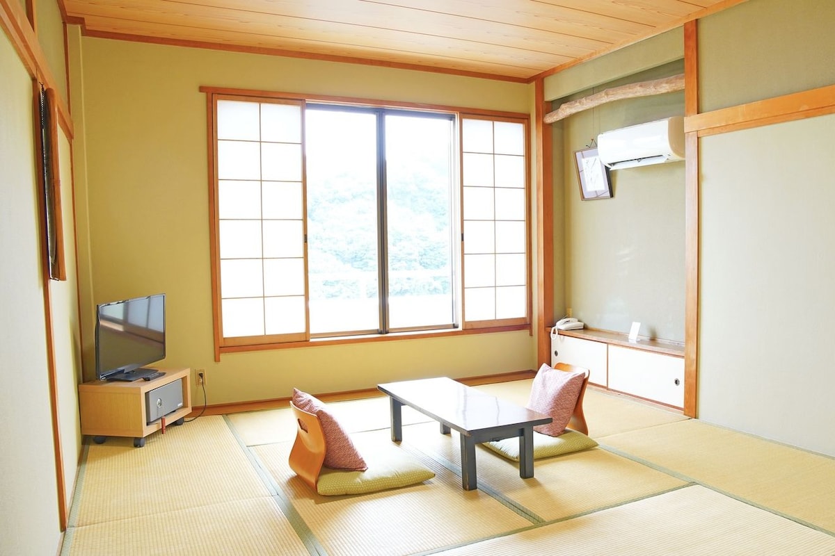 Una habitación japonesa tradicional con suelo de tatami, una mesita baja, dos cojines, un televisor, un armario pequeño y una puerta corredera.