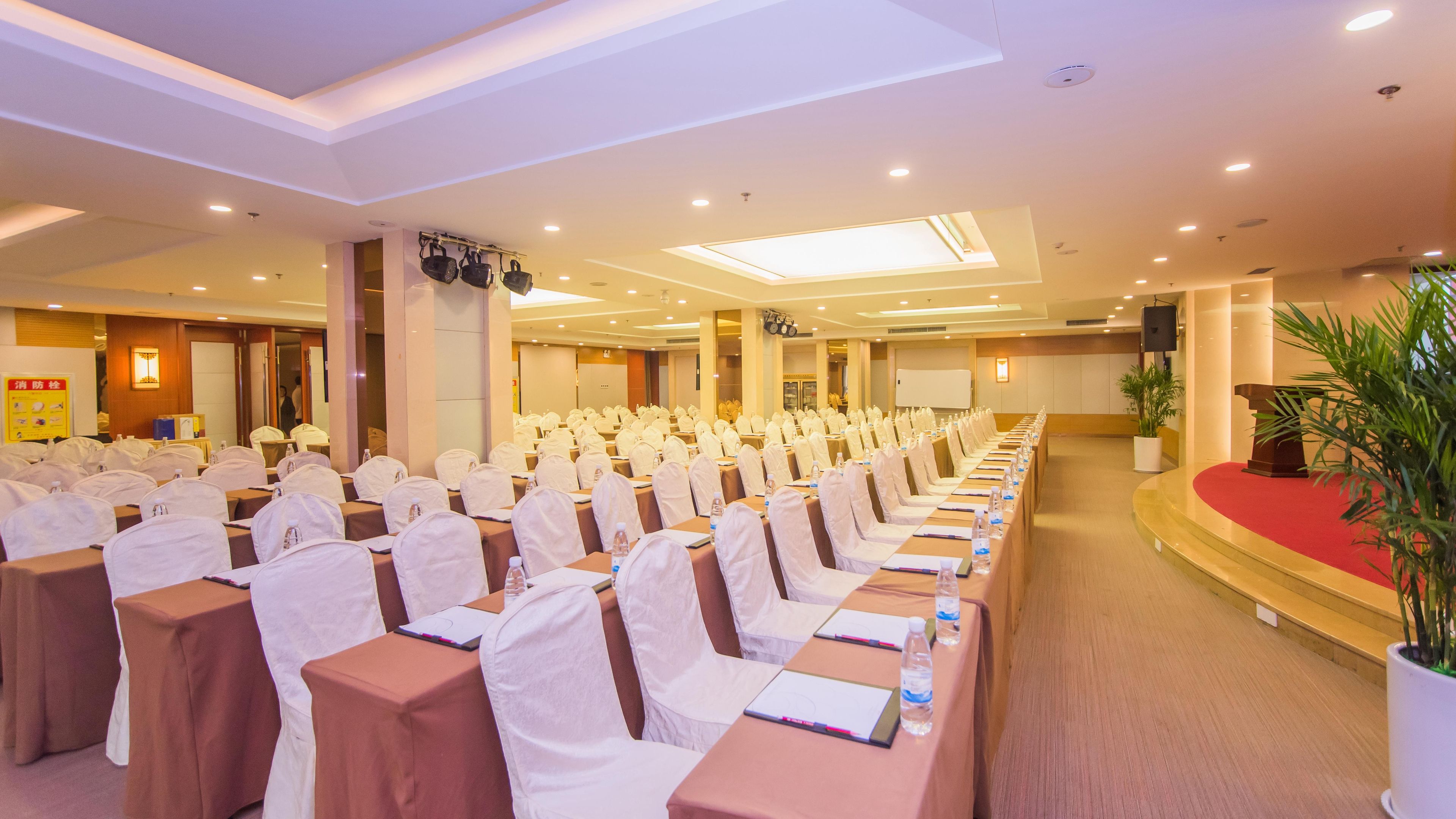 A conference room with rows of tables and chairs, a podium, and a large screen.