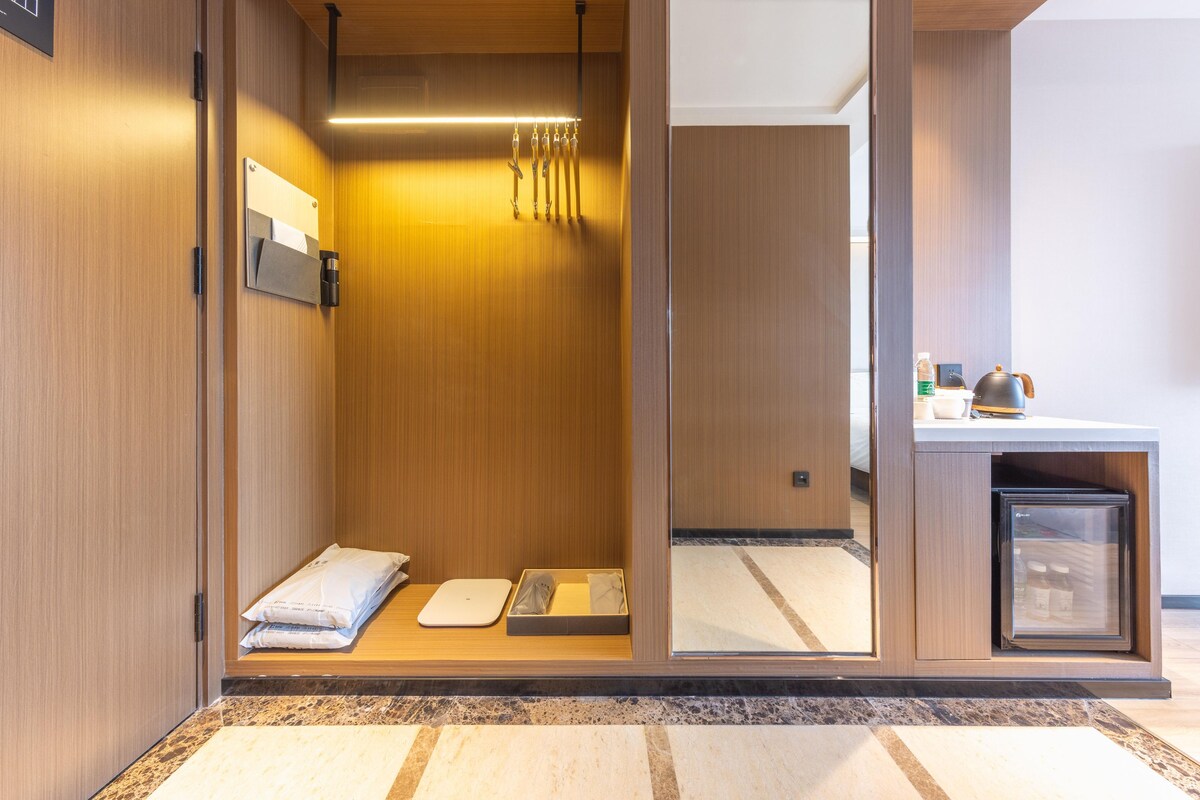 A modern hotel room with a wooden interior, a small seating area with a towel and a tray, a compact refrigerator, and a view of the adjacent room.