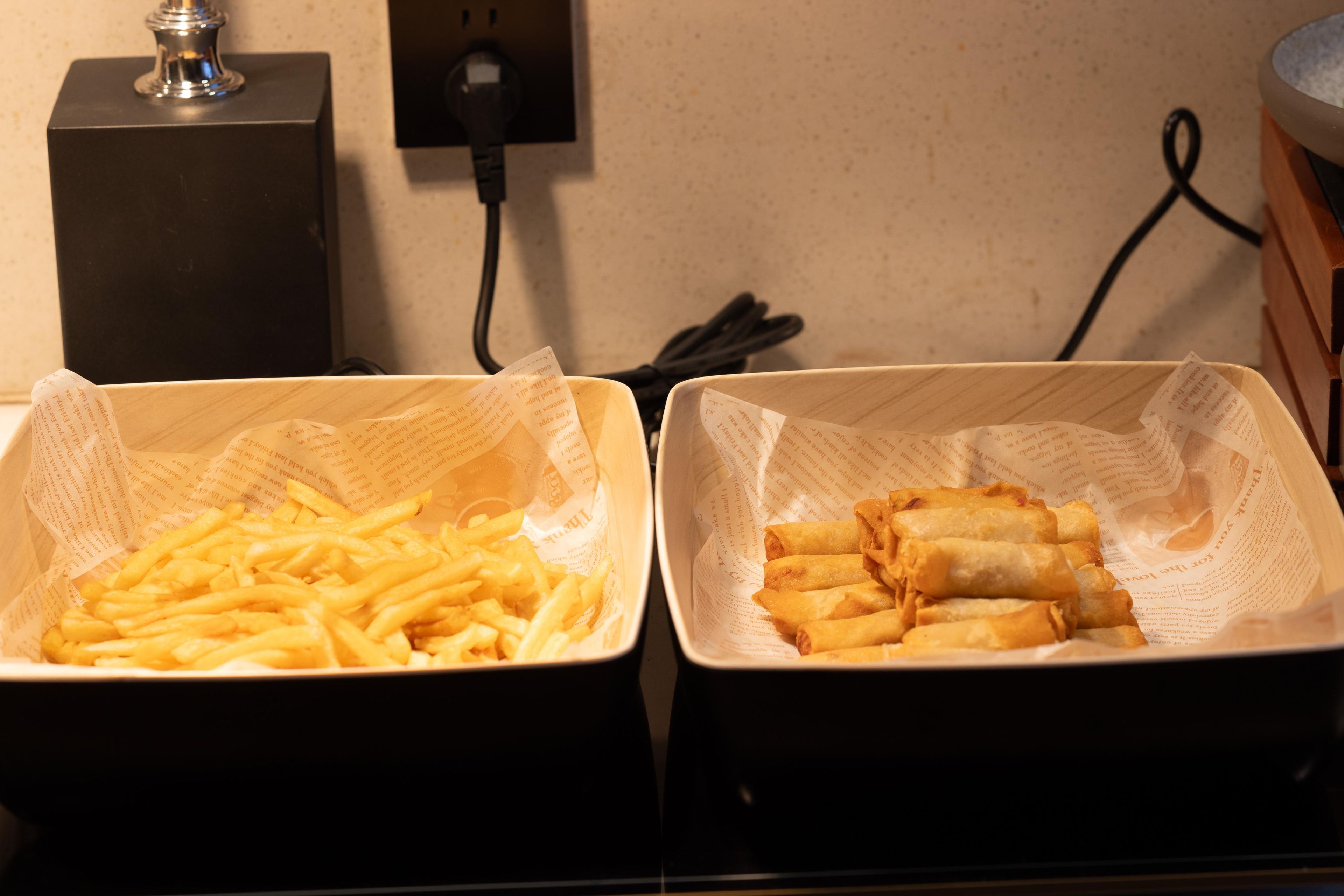 Duas bandejas de comida, uma com batatas fritas e outra com rolinhos primavera, sobre uma mesa com uma tomada e cabo elétrico ao fundo.