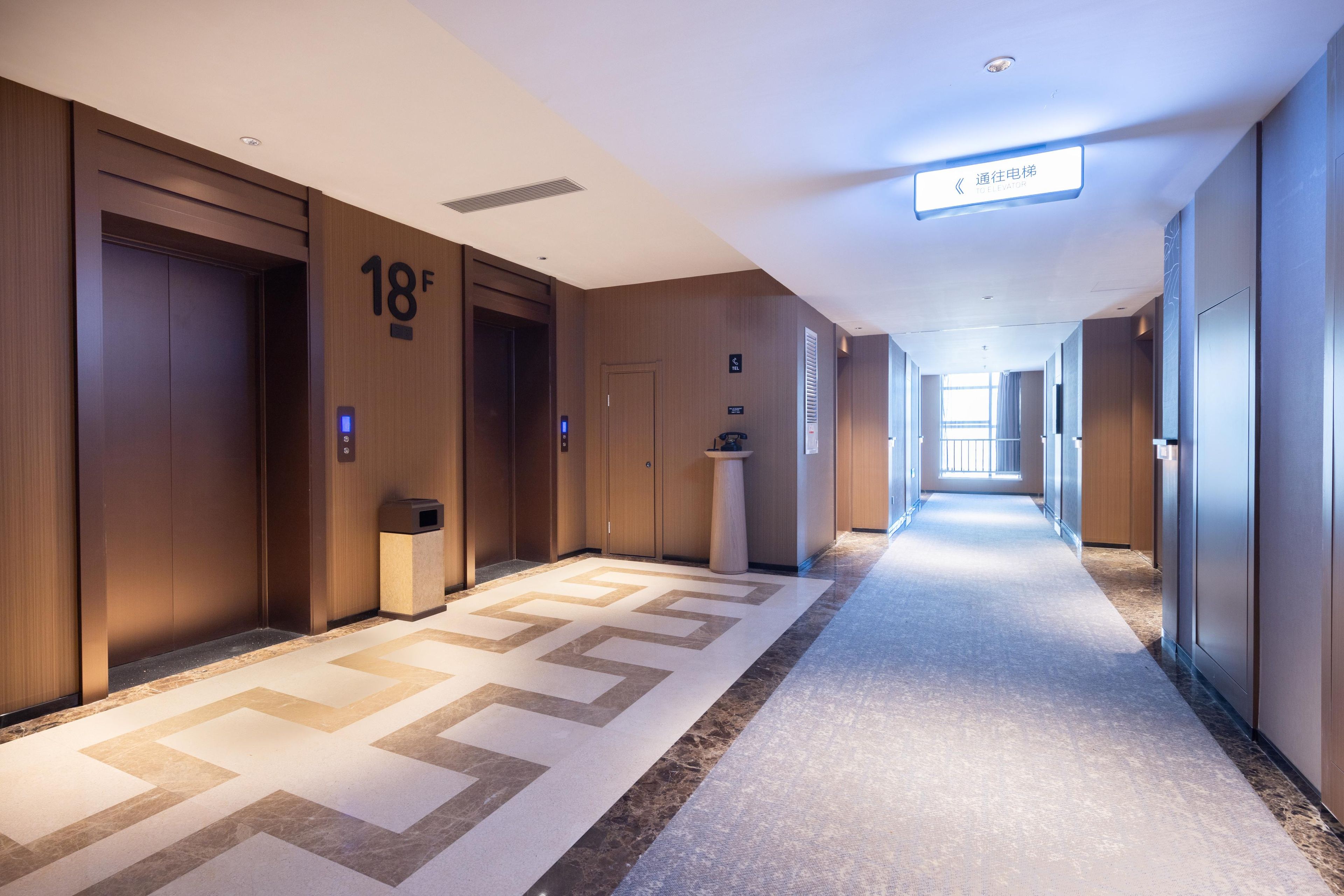 Un couloir d’hôtel avec des portes en bois, un sol recouvert de moquette et un panneau indiquant la direction de l’ascenseur.