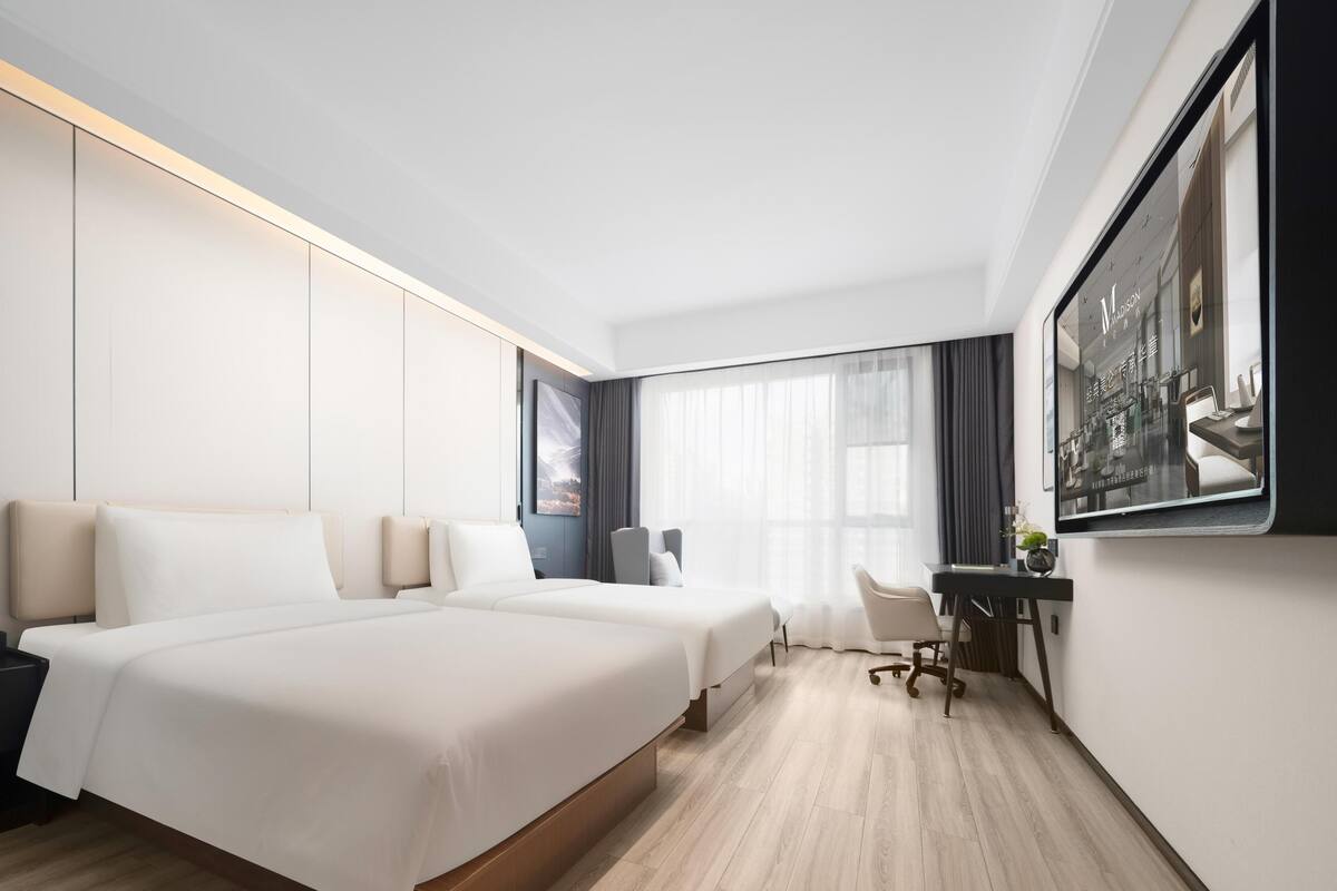 A modern hotel room with two large beds, a desk with a chair, and a large window with curtains.