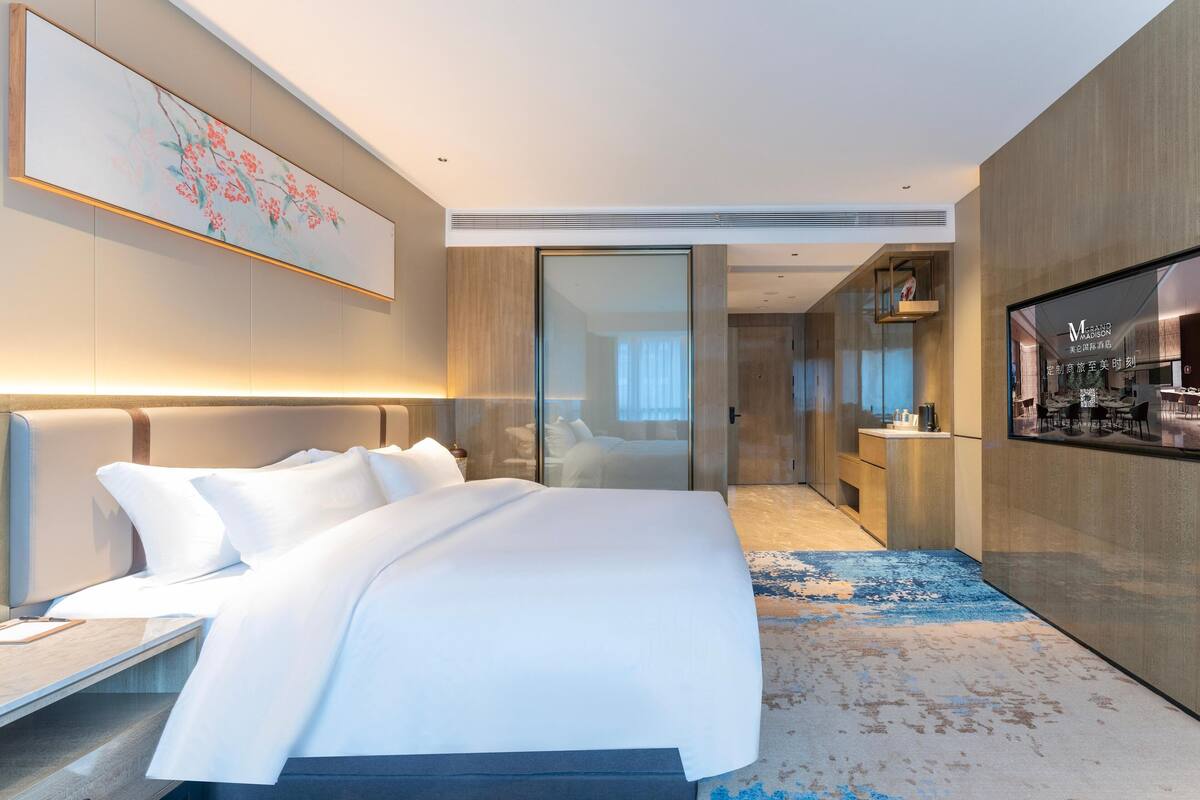 Ein modernes Hotelzimmer mit einem großen Bett, einem Schreibtisch, einem Flachbildfernseher und einem durch eine Glastür einsehbarer Badezimmer.