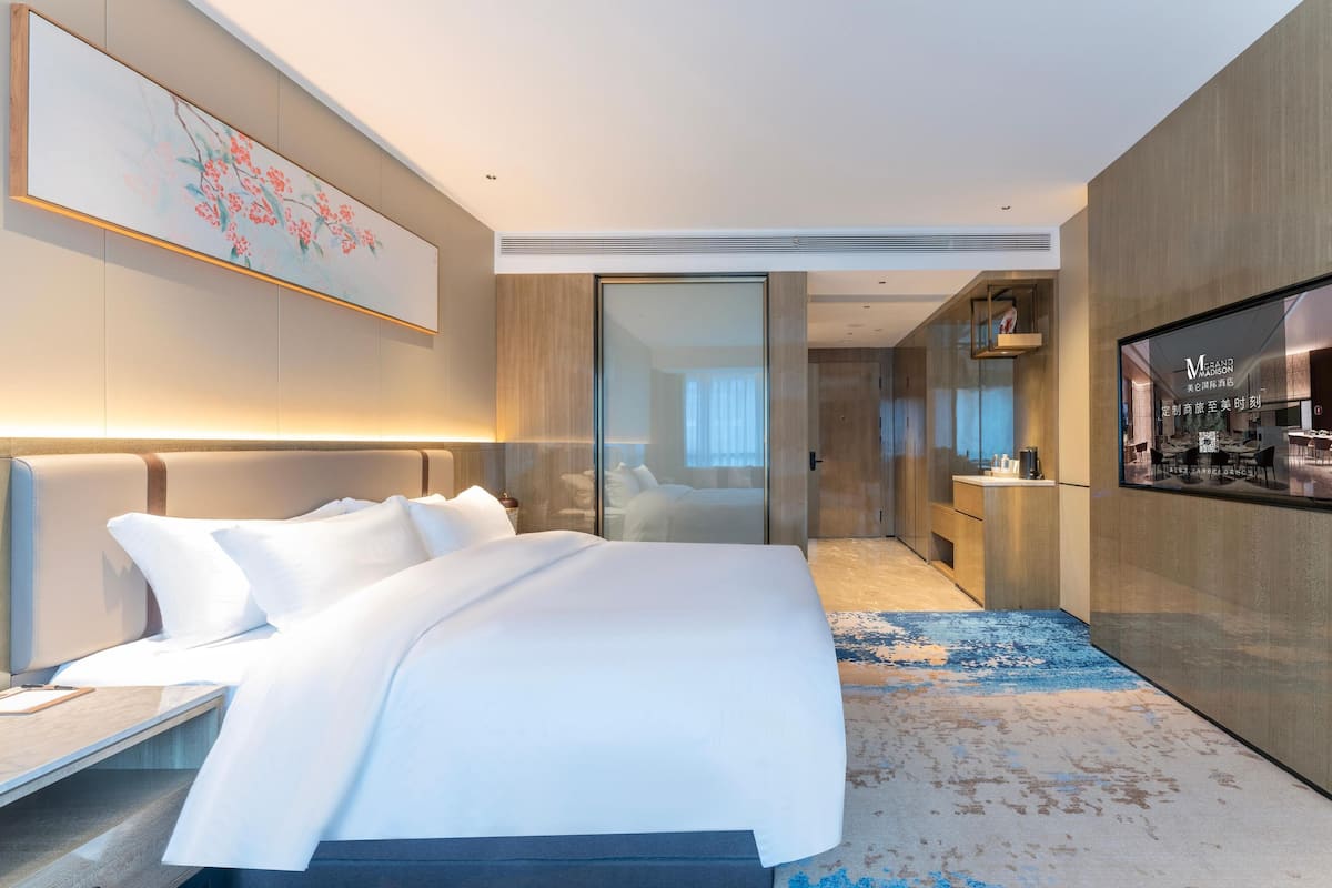 A modern hotel room with a large bed, a desk, a flat-screen TV, and a bathroom visible through a glass door.