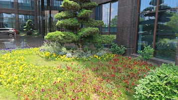 Un jardín ajardinado con diversas flores y un árbol alto frente a un edificio moderno con grandes ventanales.