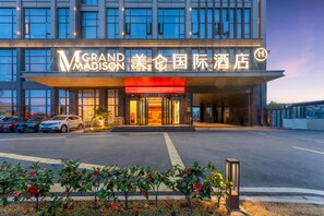 A hotel entrance with a modern facade, illuminated signage, and a parking area with several cars. - Grand Madison Hotel Quzhou High-speed Railway Sta (Quzhou)