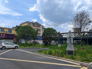 Un parking avec une statue au premier plan, un bâtiment avec une enseigne visible à l’arrière-plan et un arbre sur la droite.