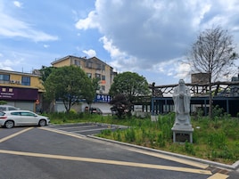 Un estacionamiento con una estatua en primer plano, un edificio con señalización al fondo y un árbol a la derecha.