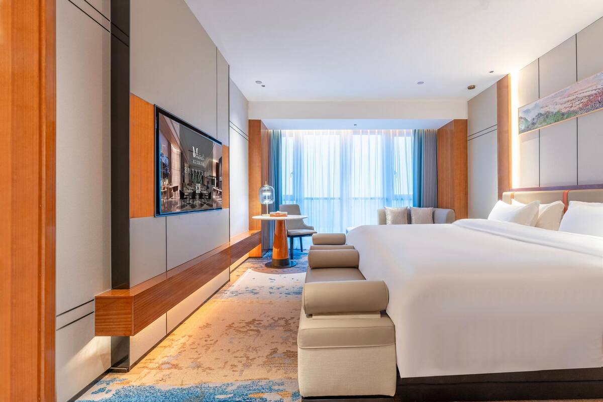 Ein modernes Hotelzimmer mit einem großen Bett, einer Sitzecke mit einem kleinen Tisch und einem großen Fenster mit Vorhängen.