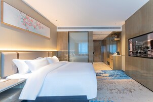 Habitación de hotel moderna con una cama grande, un escritorio, un televisor de pantalla plana y un baño visible a través de una puerta de cristal.