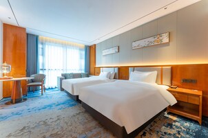 Habitación de hotel con dos camas, un escritorio, una silla y un gran ventanal con cortinas.