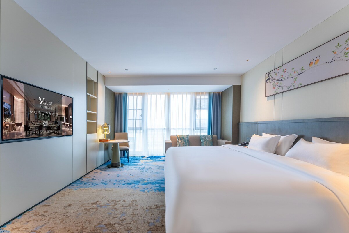 A hotel room with a large bed, a desk with a lamp, a chair, and a view of the dining area through a window.