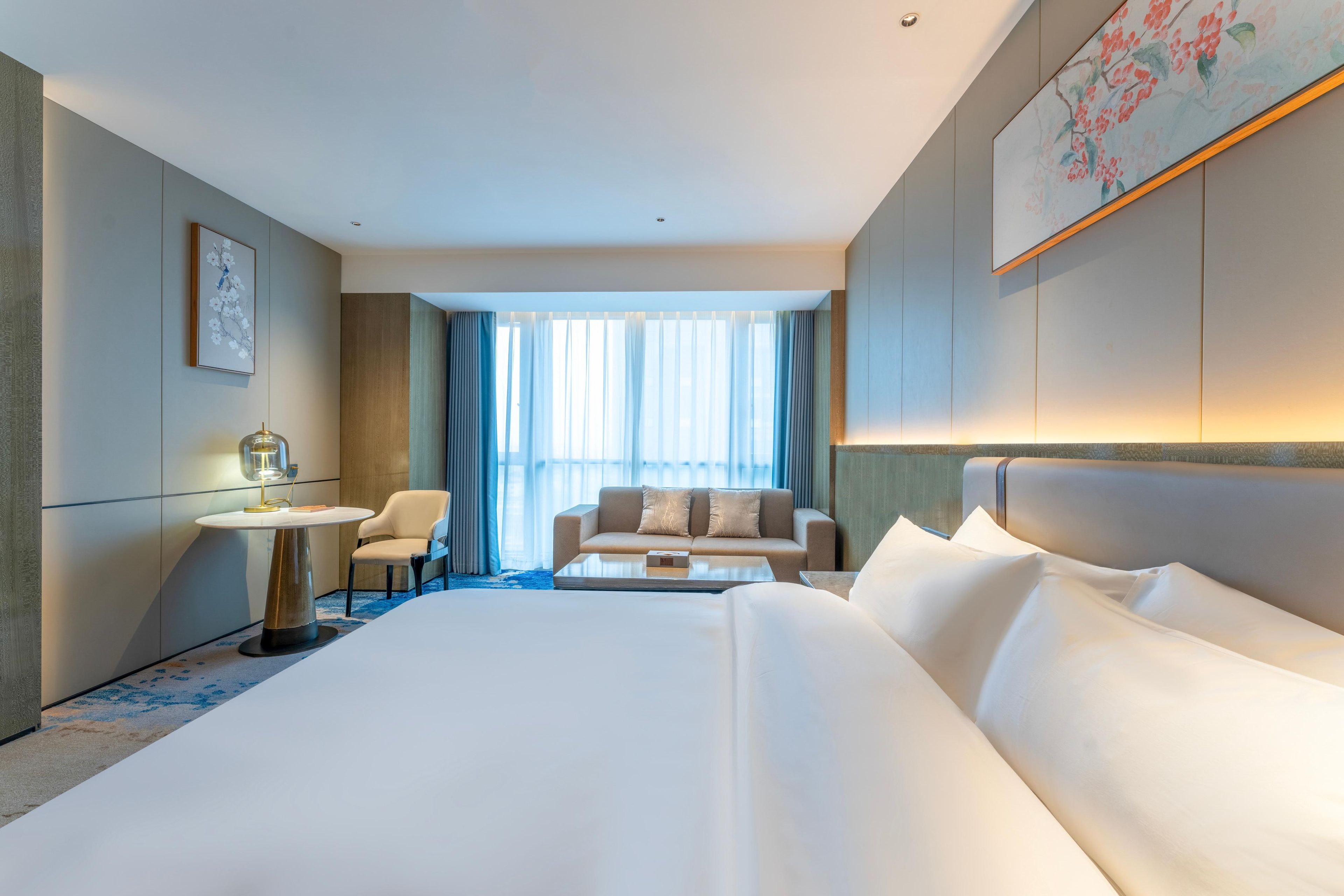 Camera d'albergo con un letto grande, un divano, un tavolino e una sedia. Si vede una finestra con tende e un quadro appeso al muro.