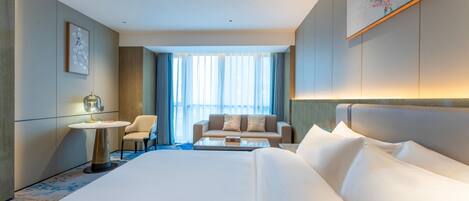 A hotel room with a large bed, a sofa, a small table, and a chair. There is a window with curtains and a painting on the wall.