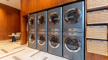 A row of four front-loading washing machines in a hotel lobby with a tiled floor and a small table with a chair.