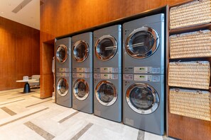 A row of four front-loading washing machines in a hotel lobby with a tiled floor and a small table with a chair. - Grand Madison Hotel Quzhou High-speed Railway Sta (Quzhou)