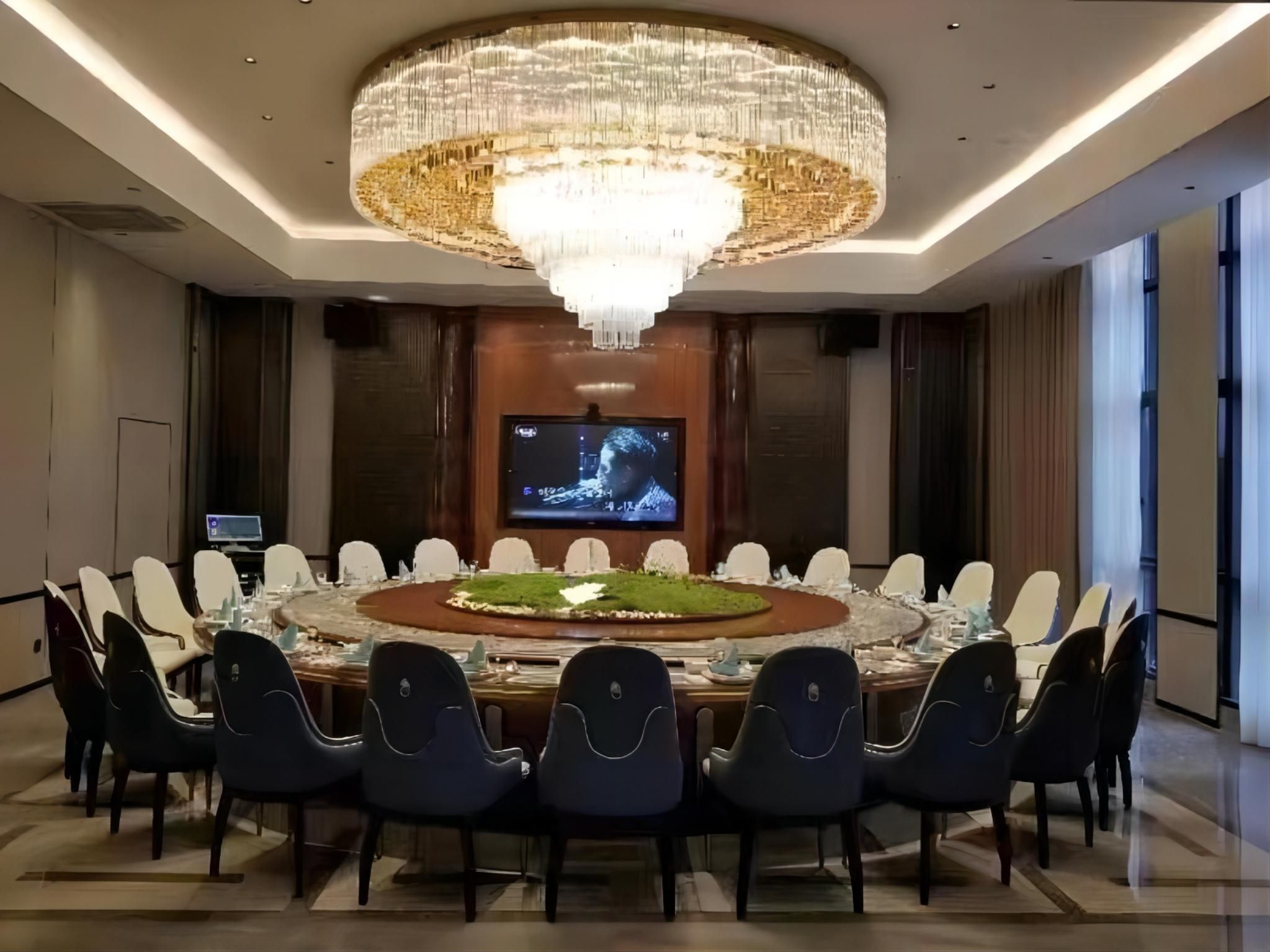 Un salón de conferencias con una gran mesa redonda, varias sillas, un candelabro, una pantalla de televisión y una pantalla pequeña sobre la mesa.