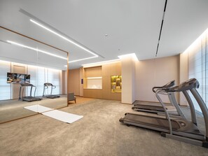 Une salle de sport moderne équipée de tapis de course, d’un tapis de yoga et d’un espace d’accueil avec un téléviseur diffusant des images d’une chambre.