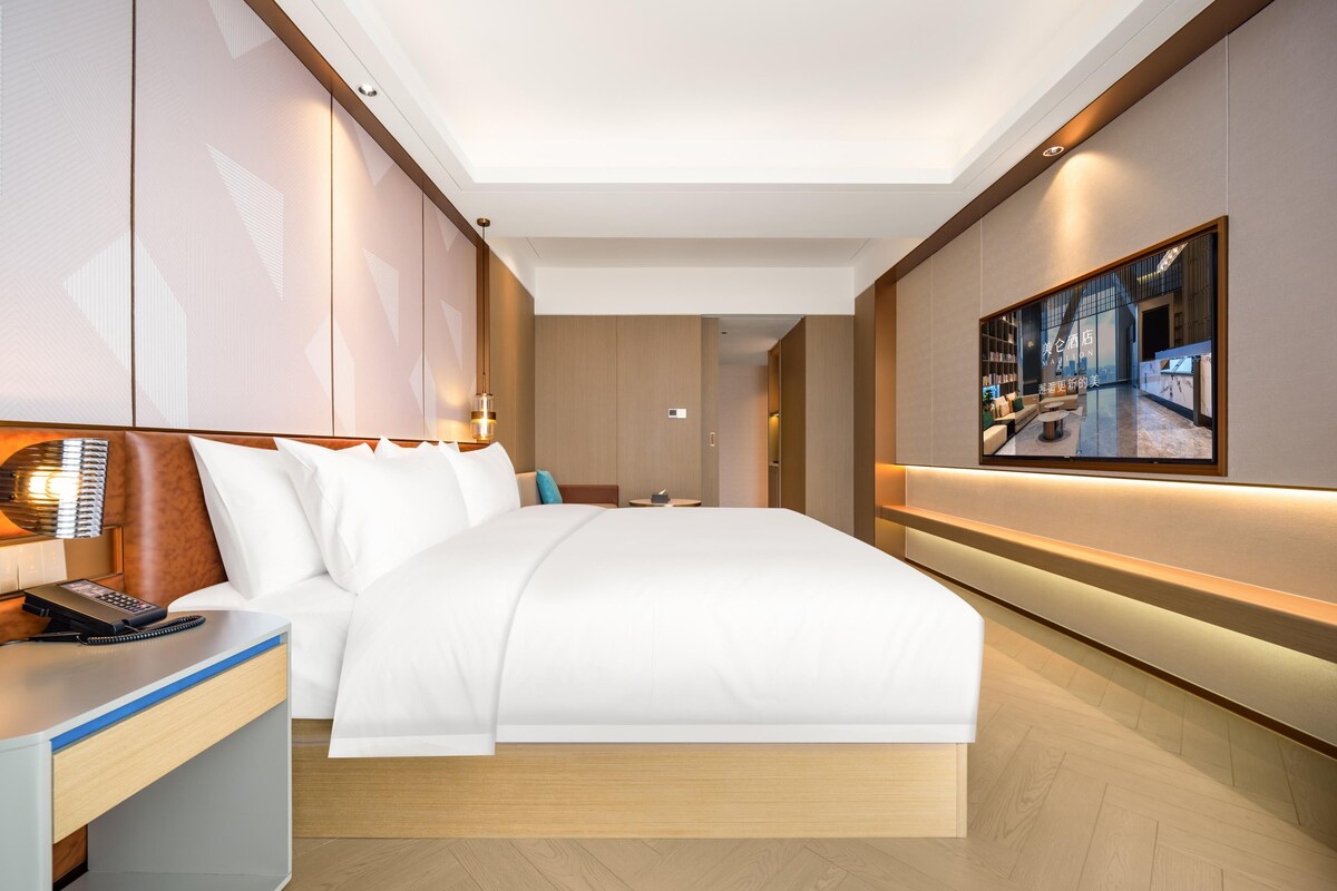 A modern hotel room with a large bed, a bedside table with a phone, a wall-mounted TV, and a hallway leading to another room.