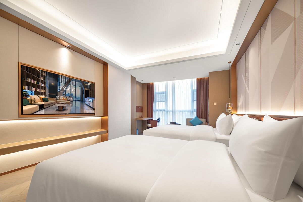 Habitación de hotel con una cama grande, zona de estar con sofá y sillones, una mesita y un ventanal con cortinas.