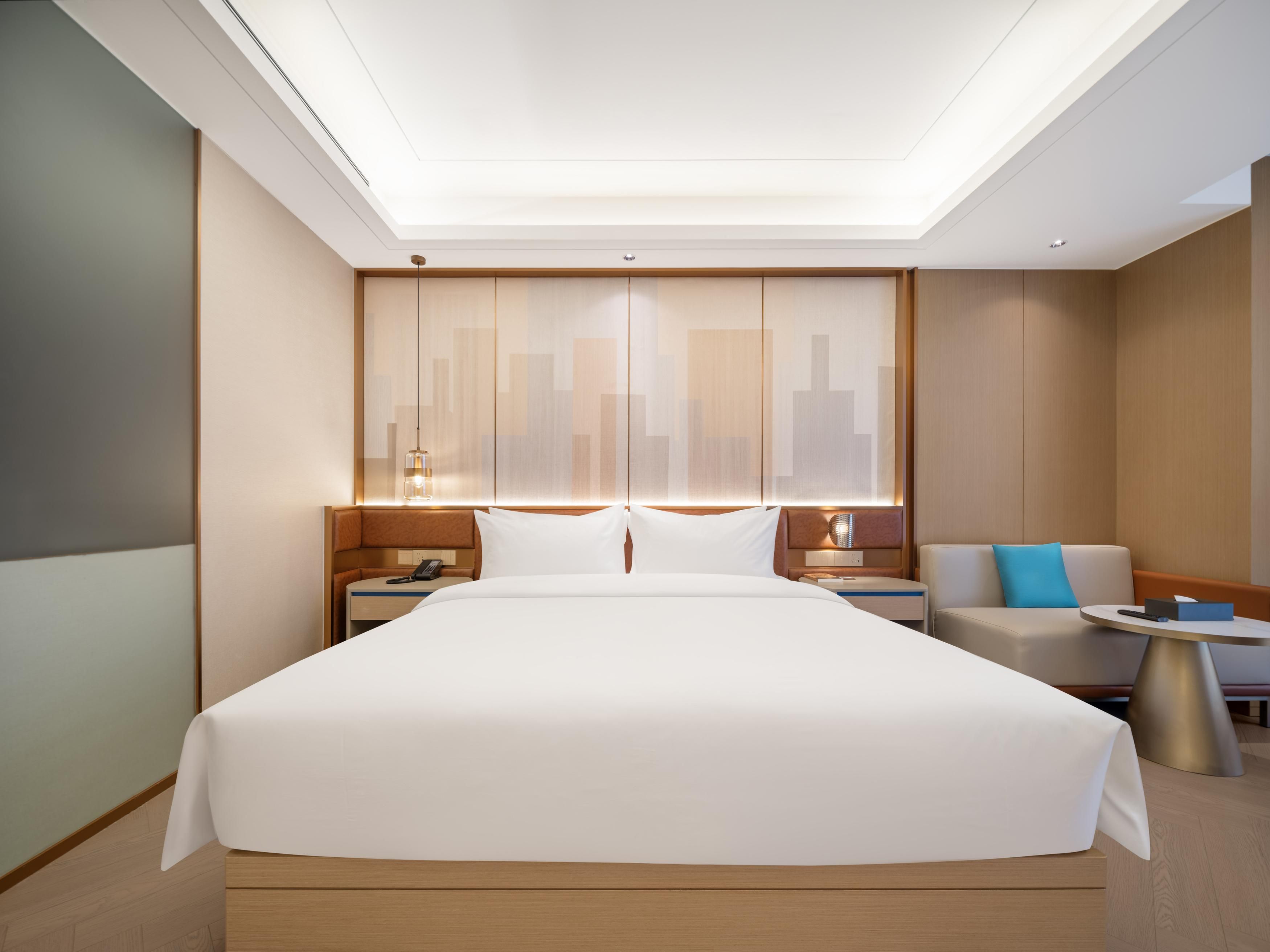 A modern hotel room with a large bed, a sofa, a small table, and a cityscape view through the window.