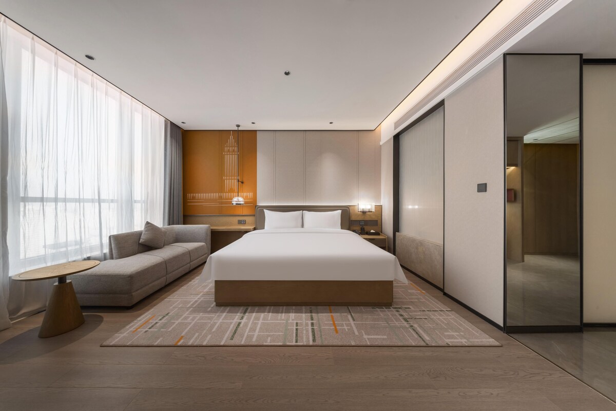 Una habitación de hotel moderna con una cama grande, un sofá, una mesita de noche y un ventanal con cortinas translúcidas.
