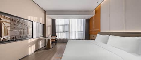 A modern hotel room with a large bed, a desk, and a window with sheer curtains.