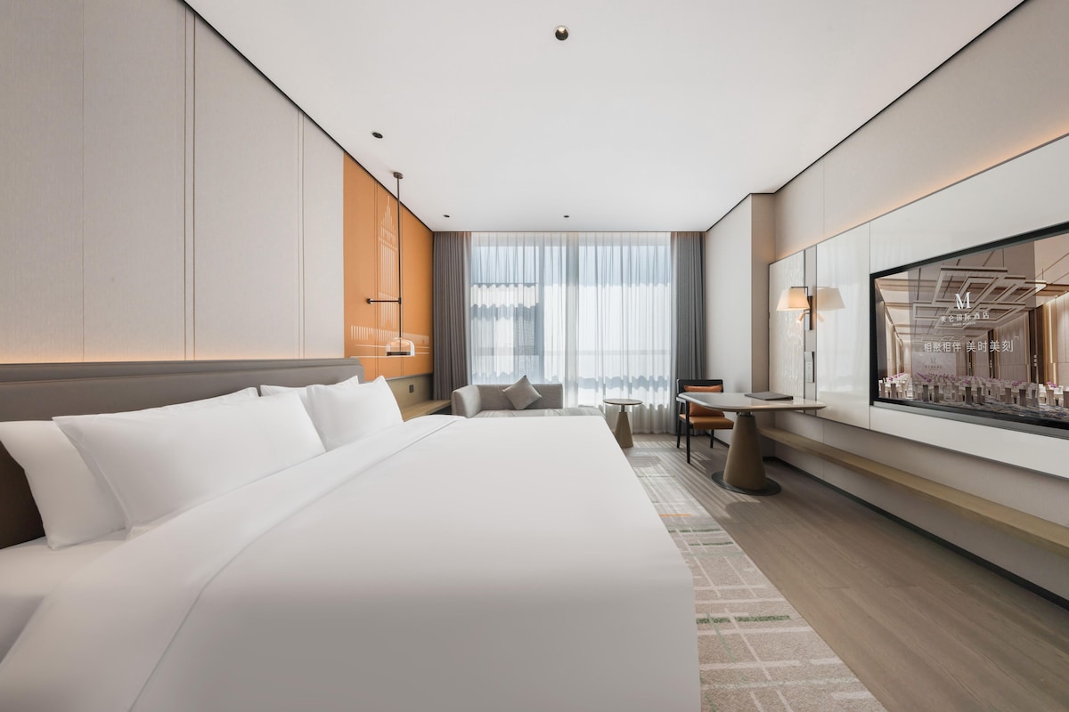 A modern hotel room with a large bed, a seating area, a small table, and a large window with curtains.
