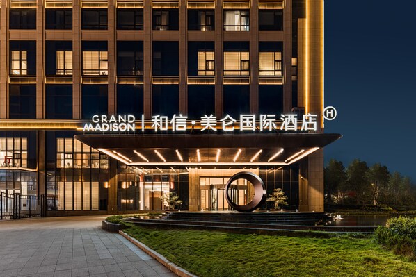 Entrada de hotel con señalización iluminada, una escultura circular y un jardín paisajista.