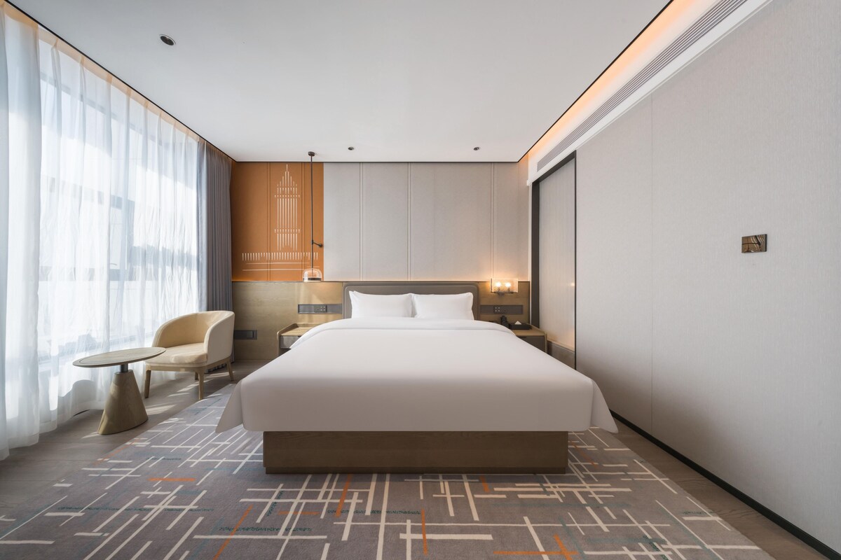 Una habitación de hotel moderna con una cama grande, una zona de estar con una silla y una mesita, y un ventanal con cortinas translúcidas.
