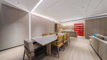 A modern dining area with a long table and chairs, a kitchen in the background, and a red storage unit.