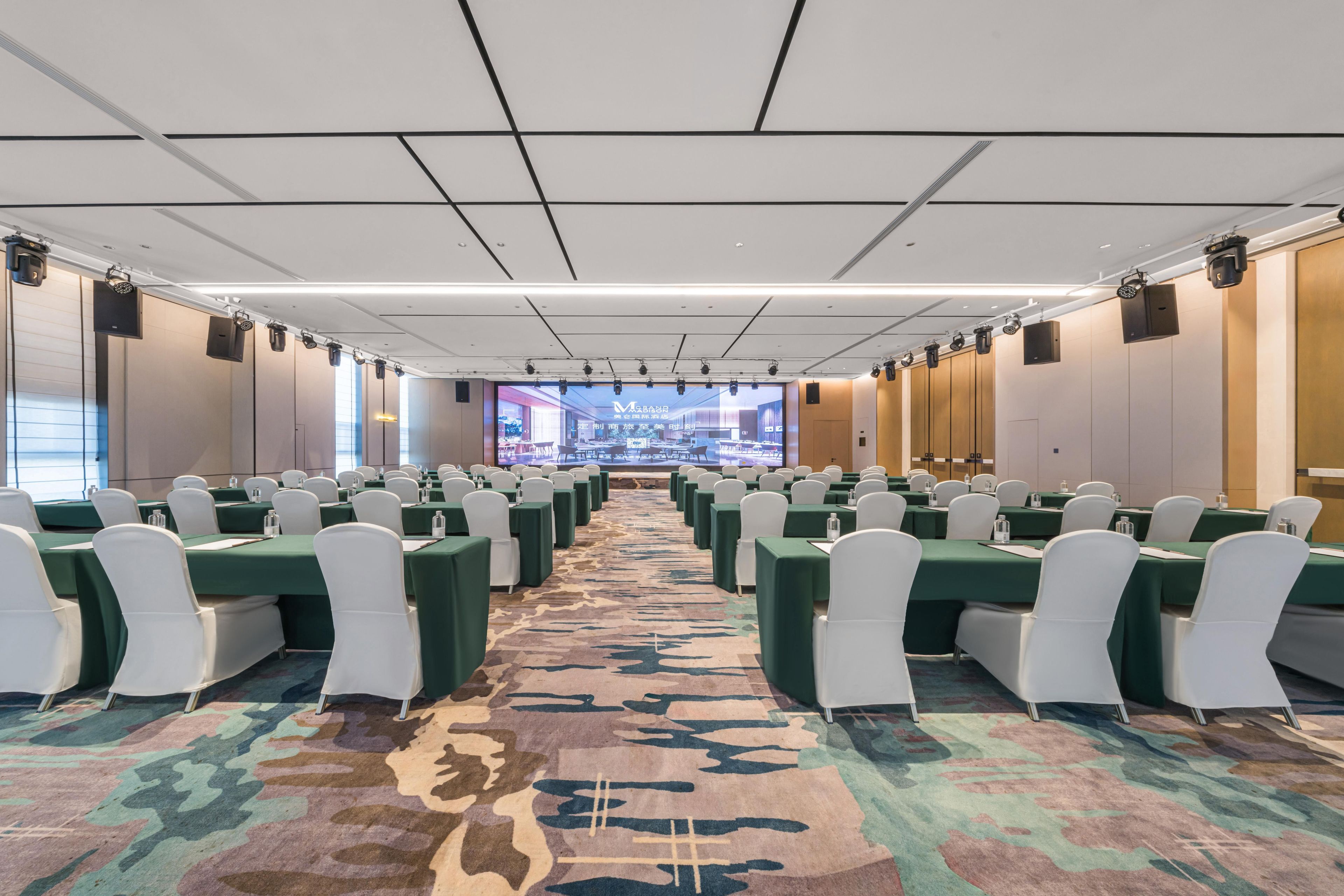 Une salle de conférence avec des rangées de tables et de chaises, un grand écran à l’avant et une moquette à motifs.