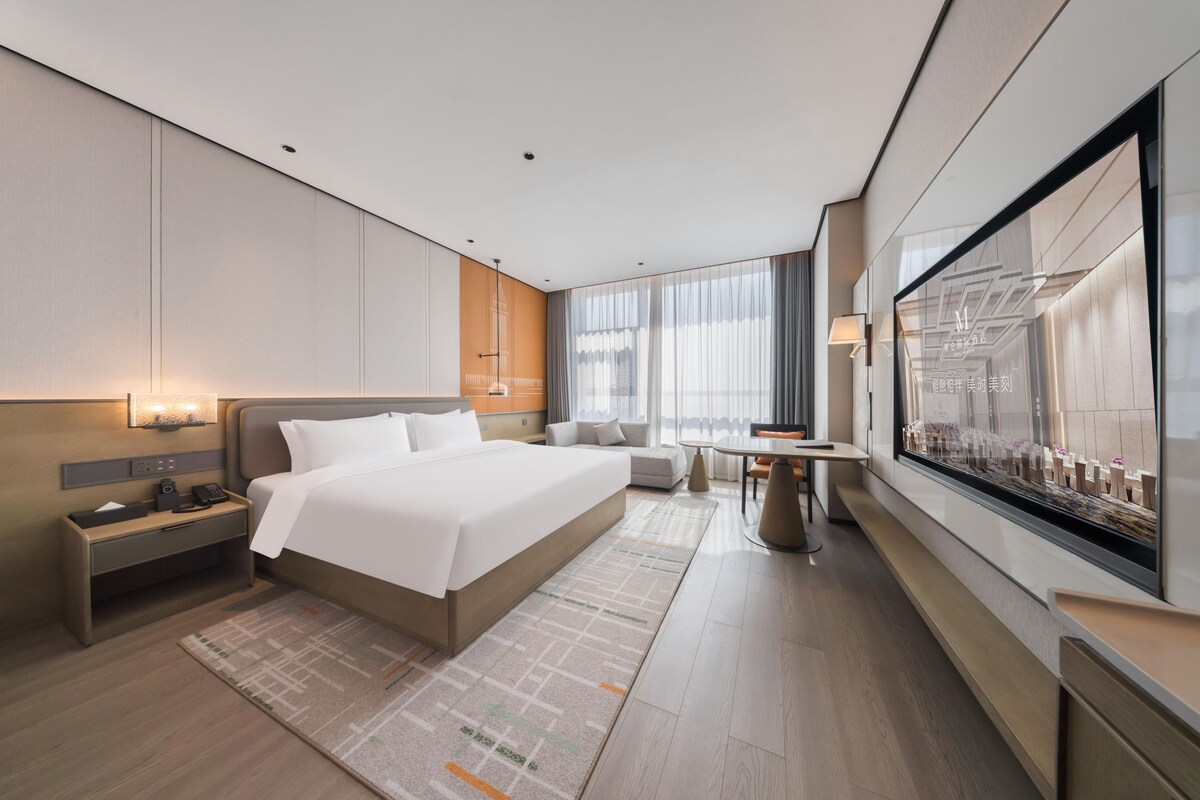 A modern hotel room with a large bed, bedside tables, a seating area, and a large window.