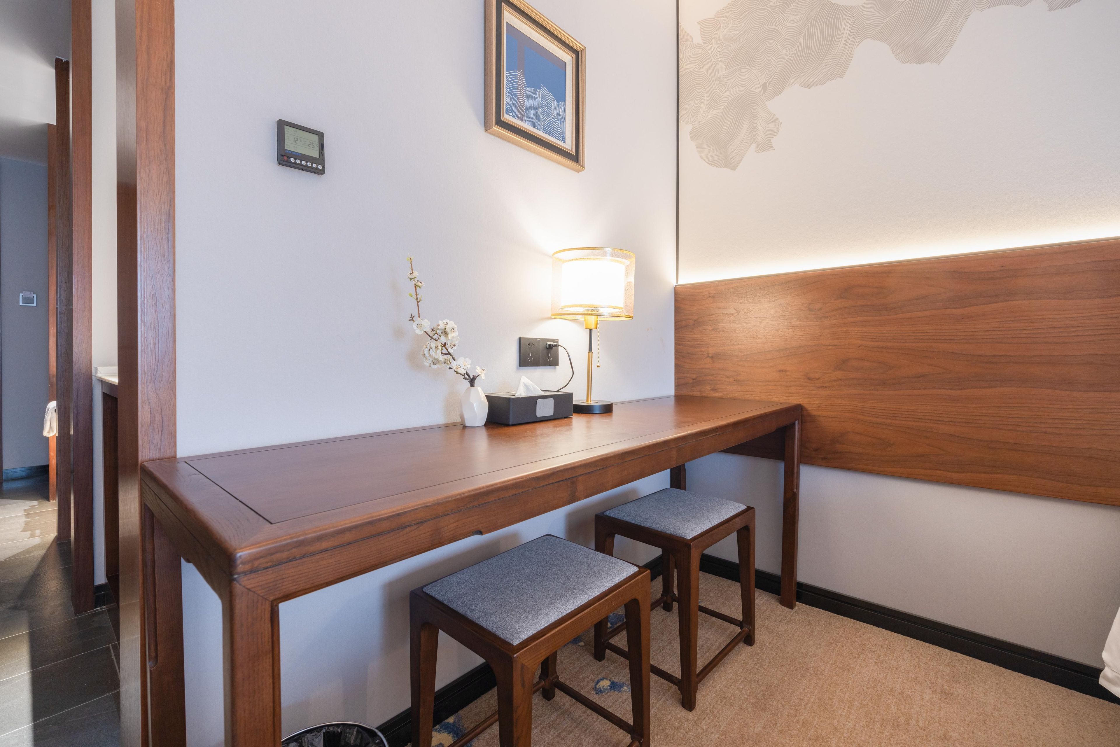 Ein Hotelzimmer mit einem Holztisch, zwei passenden Stühlen, einer Lampe, einem gerahmten Bild und einem Thermostat an der Wand.