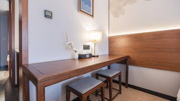 A hotel room with a wooden desk, two matching chairs, a lamp, a framed picture, and a thermostat on the wall.