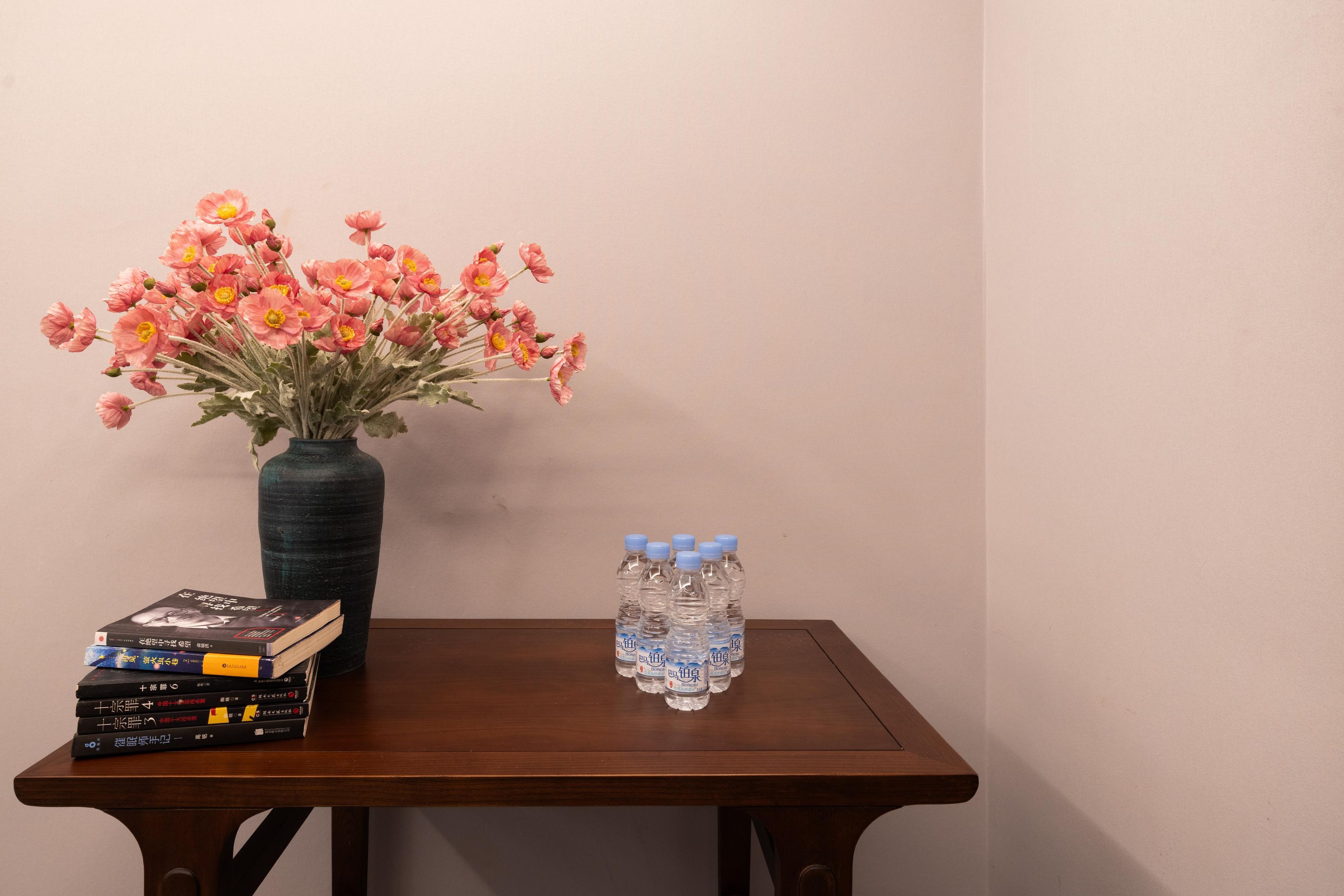 A wooden table with a vase of pink flowers, a stack of books, and six water bottles.
