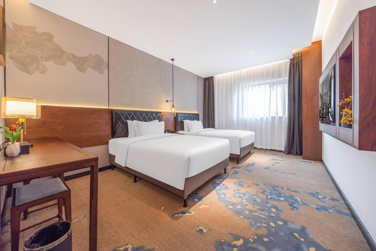 A hotel room with two beds, a desk, a trash bin, and a large window with curtains.