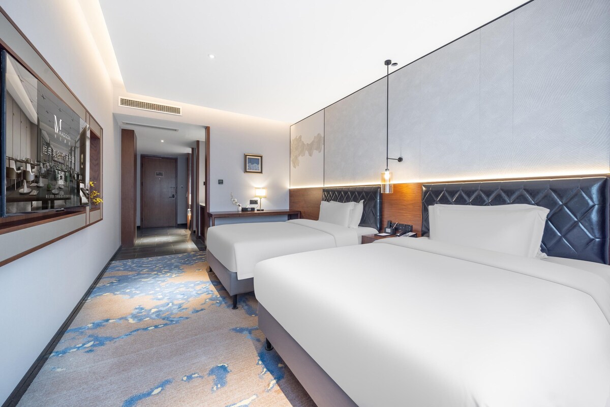 A hotel room with two beds, a large mirror, a desk with a lamp, and a view of the hallway.