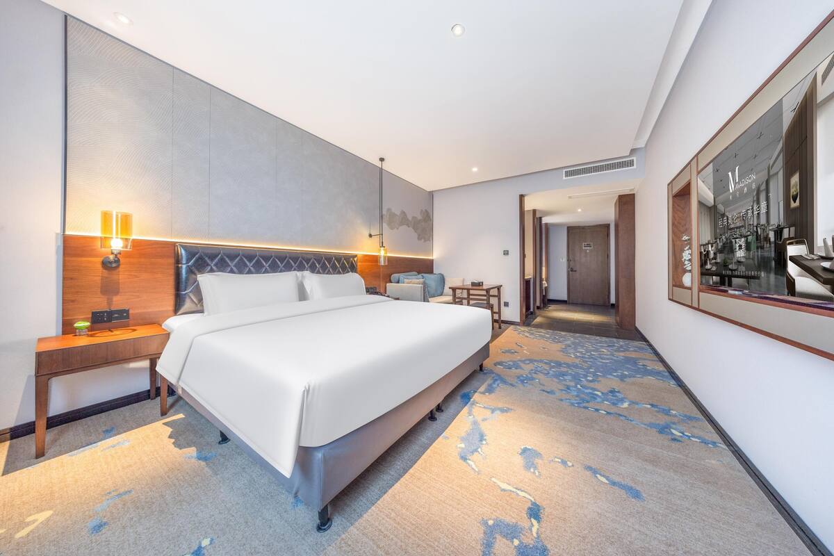 A modern hotel room with a large bed, bedside lamps, a small table, and a view of the hotel lobby.