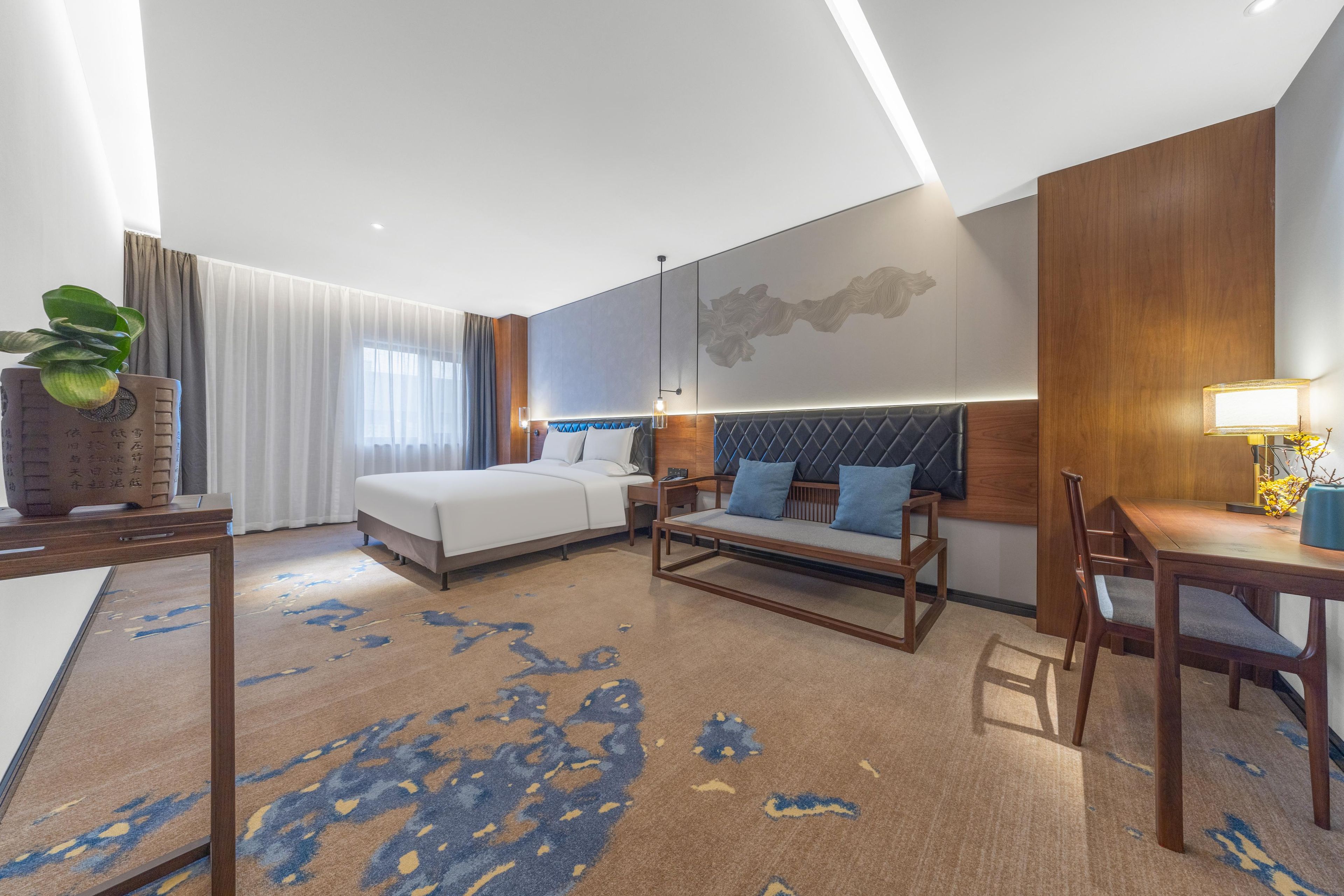 A modern hotel room with a bed, sofa, dining table, and a large window with curtains.