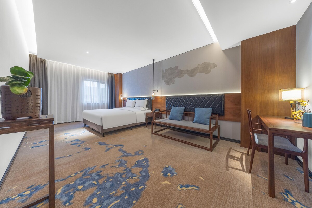 A modern hotel room with a bed, sofa, dining table, and a large window with curtains.