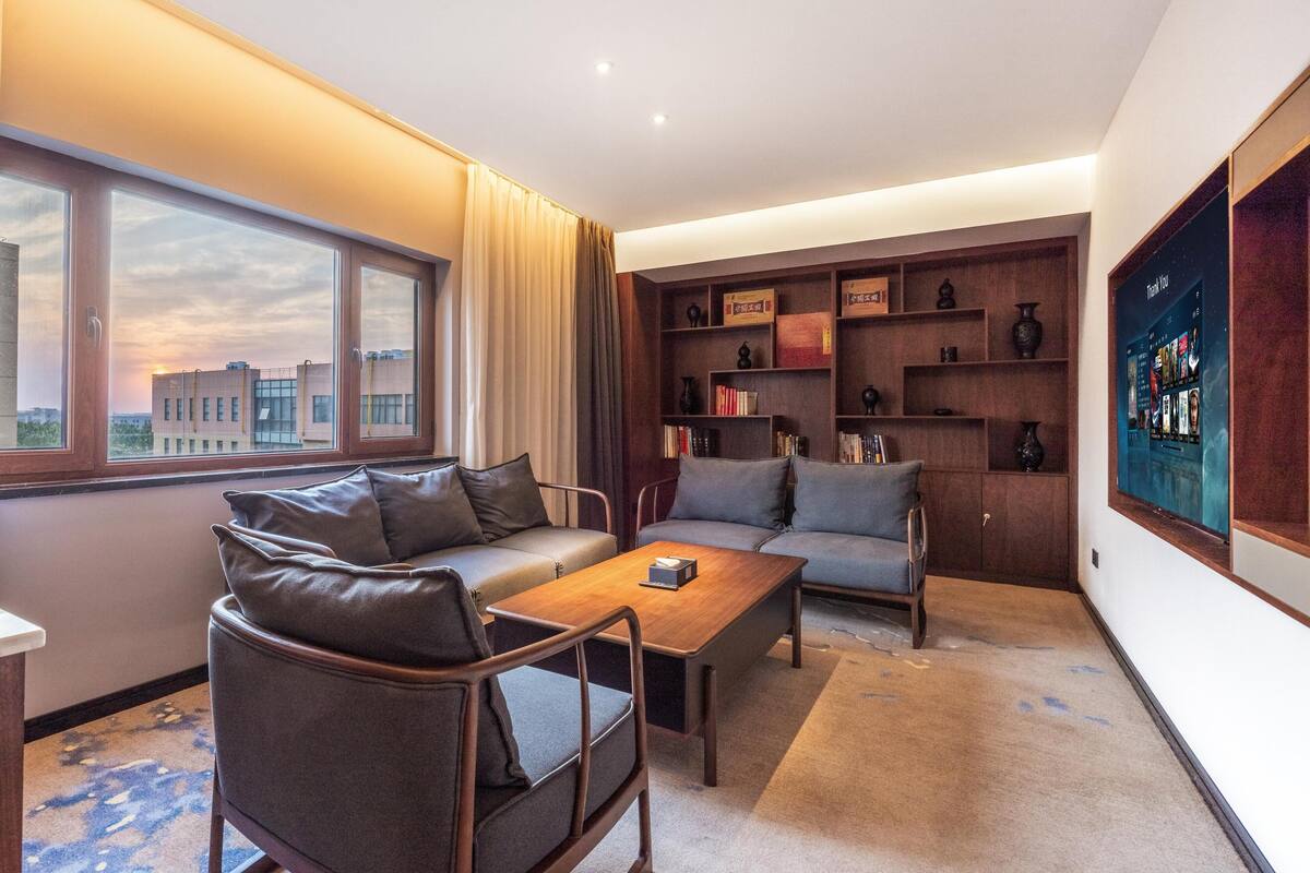 A modern living room with a sofa, armchair, and coffee table. There is a bookshelf with books and decorative items. A large window offers a view of buildings outside.