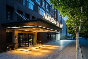 Das Madison Hotel mit moderner Fassade, beleuchtetem Eingang und einem von Bäumen gesäumten Weg.