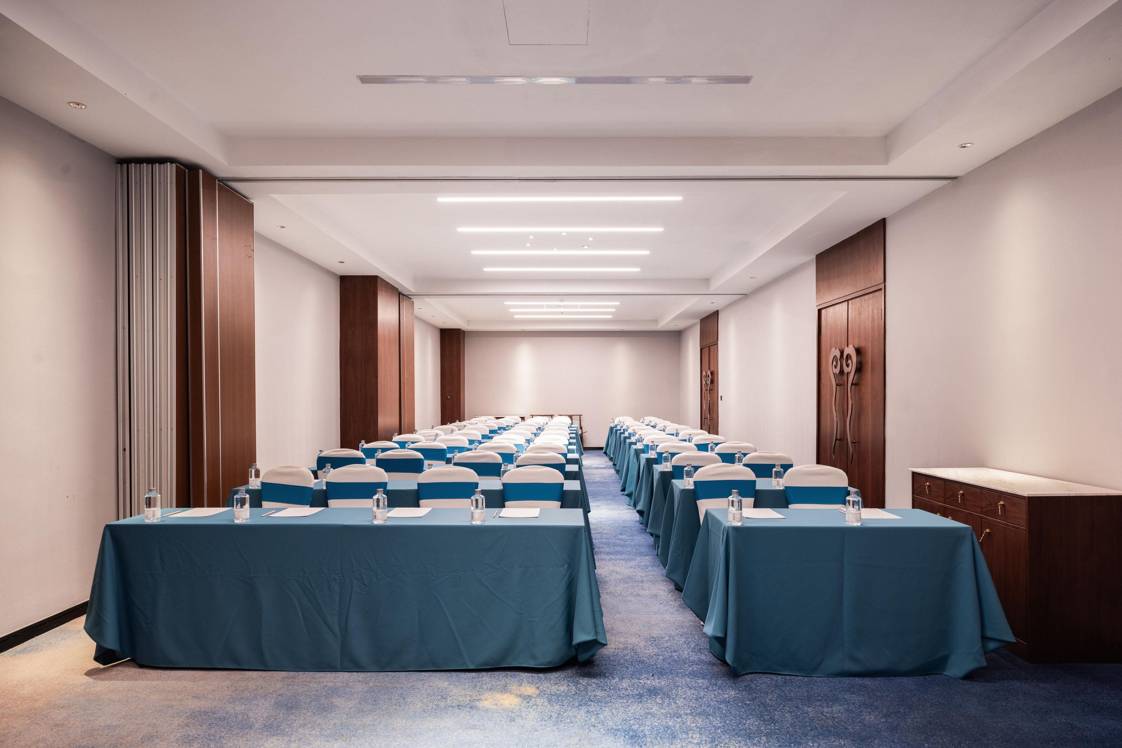 A conference room with rows of tables, each with a blue tablecloth and white chairs, equipped with water bottles and notepads.